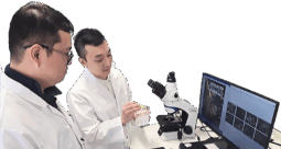 Two scientists in white lab coats looking at a material sample in front of a microscope