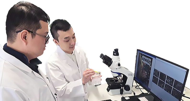 Two scientists in white lab coats looking at a material sample in front of a microscope