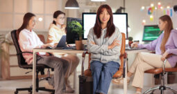 Young looking woman sitting cross-armed and smiling with other young women in the background working on computers