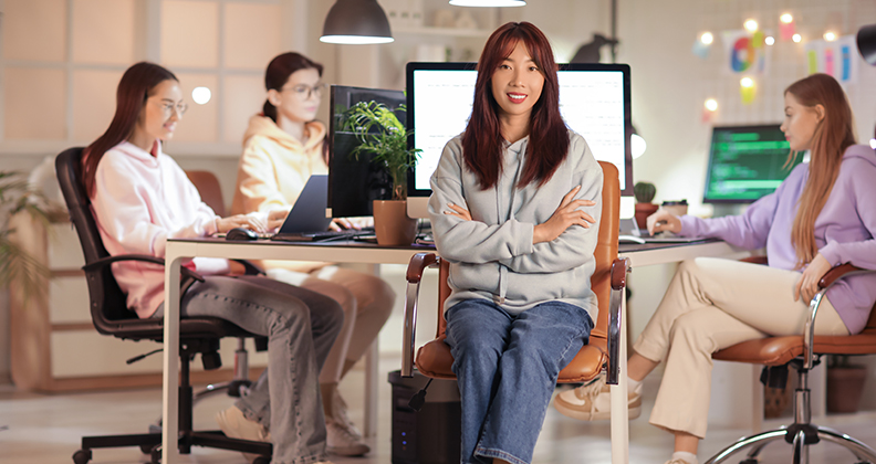 Young looking woman sitting cross-armed and smiling with other young women in the background working on computers