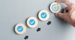 Checkmarks in wooden circles ascending to a target graphic held by a hand