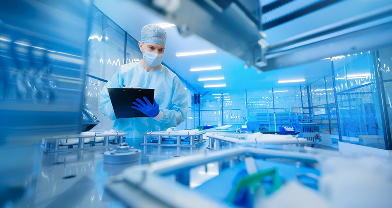 Masked and gowned worker holding a clipboard and looking at machinery
